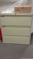 Front view of the beige three-drawer filing cabinet with a small box on top, showing smooth beige finish and drawer handles.
