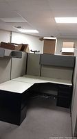 View of L-shaped office desk with filing cabinet drawers under desk, and attached fabric-covered cubicle panels in light neutral color.