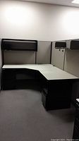 Full office cubicle set showing L-shaped desk, overhead shelf, fabric-covered panels with black framing, and under-desk filing cabinets.