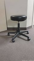 Full view of the black rolling stool showing round cushioned seat, adjustable post, and five caster wheel base.