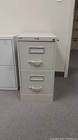 Front view of white thin 2 drawer filing cabinet standing on carpeted floor next to another filing cabinet. Shows handles and label holders on each drawer.
