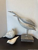 Group photo showing two tall gray wooden bird sculptures on wooden bases and two smaller ceramic bird figurines in front, one white and one speckled.