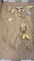 Multiple gold coloured brooches and necklaces laid out on cardboard showing detailed shapes and motifs including leaves, animals, apple, acorn, and pendants on chain necklaces.