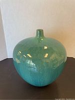 Front view of the large rounded ceramic pot with narrow neck in blue-green glaze on black surface against white backdrop.