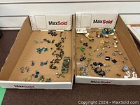 Two trays showing assorted costume earrings and necklaces spread inside cardboard boxes.