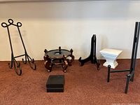 Set of five decor stands on carpeted floor: black metal easel, wooden round display stand, small black candle holder, white wooden pedestal with metal frame, tall black metal stand with scroll design