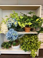 Wide view of white and blue faux hydrangea flowers, multiple faux ivy plants, and other green faux foliage on shelving
