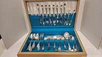 Flatware set inside open wooden box showing spoons, forks, knives and other utensils arranged in rows inside blue felt-lined bottom and white satin-lined lid.