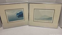 Two framed landscape prints positioned side by side, showing blue and gray mountain scenes with white mats and silver frames.