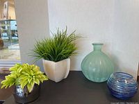 Green glass vase, silver metal pot, white ceramic pot with faux plant on table against a light background.