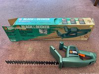Black & Decker electric hedge trimmer positioned in front of its original cardboard box showing product image and branding.