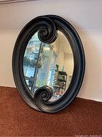 Oval mirror with black frame and scroll detail at top and bottom, showing reflective glass, photographed on a carpeted floor near a white wall.