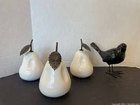 Three white ceramic pears with metal stems and leaves arranged on a white background and black bird figurine standing beside them.