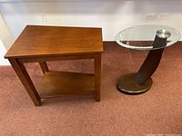 Wood rectangular side table and oval glass side table shown side-by-side on carpeted floor against white wall.