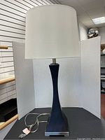 Full view of the contemporary tall table lamp with white drum shade and tapered dark square base on a black surface.