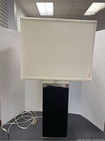 Front view of square table lamp showing white fabric shade and black base with metal trim, with power cord laid out to the side