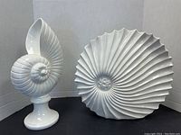 Two white ceramic shell decor pieces against white background; spiral shell on pedestal and large ridged sunburst shell