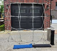 Metal foldable frame expanded and set up in front of garage door with one blue and one black carry bag on ground and collapsed frame segments next to it.