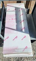 Stack of four white polycarbonate sheets covered with protective film, stacked on a metal surface outdoors in sunlight.
