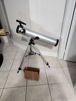 Front side view of the silver telescope mounted on the adjustable tripod, with a cardboard box on the floor below.