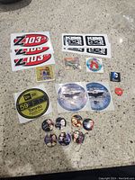 Photo of various branded stickers and promotional buttons including Z103.5, Kiss 92 FM, Pokémon, Digimon, and DC Comics branding, laid out on a countertop.
