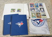 Image showing binder with baseball cards and two blue spiral notebooks with Blue Jays logo, white cloth with Blue Jays logo placed underneath.