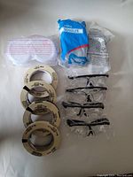 Photo showing five rolls of 3M masking tape, three pairs of white safety goggles, and five pairs of black safety glasses wrapped in plastic.
