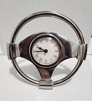 Front view of the racing wheel shaped clock showing the circular metal frame and the clock face with Roman numerals.