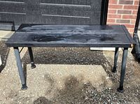 View of a metal work bench with a solid rectangular metal top and four tubular legs on uneven ground outdoors against a brick wall.