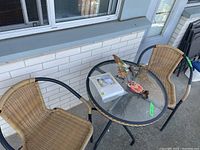 Overview of entire bistro set including glass table, two wicker chairs, new patio chair cover still wrapped, and metal wall decor on table.