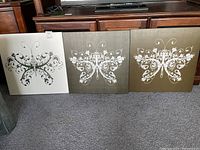 Three square canvas prints featuring decorative butterfly designs in white floral and ornamental patterns on neutral backgrounds, arranged side by side on carpet.