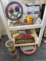 Shelf with stained glass floral decorative piece, assorted candles including pillar candles and boxed candles, candle holder trays, and tablecloth clips.