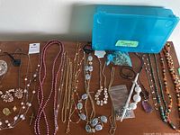 Photo of assorted costume jewelry items on a wooden surface including beaded and chain necklaces, an earring holder, and a ring dish with a blue plastic container in the background