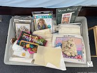 Image showing several cross stitch kits by Dimensions, Royal Paris, Bow Ties, and Door Belles along with fabric pieces and an assortment of embroidery supplies in a plastic bin.