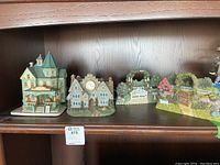 Full shelf view with all four items arranged side by side: Victorian Bed and Breakfast figurine, resin clock building, and two arbour wall hangings with greenery and white fences.