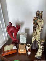 View of the entire lot showing all items on a wooden surface including wooden cat statue, ceramic plinth, photo frame music box, pottery vases, painted vessels, and figurine statues.