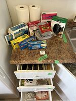 Kitchen countertop with various kitchen supplies including plastic bags, foil, burner liners, paper towels, and drawer partially open showing cutlery and towels.
