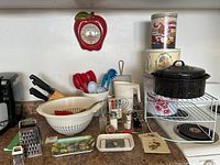 Full overview of kitchen items showing apple clock, utensil block with knives, strainers, roasting pot on wire shelf, salt and pepper shakers, tin cans, trays, and graters.