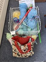 Top view of the wire basket filled with yarn balls, knitting needles, knitting pattern, and part of tote bag.