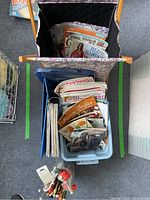 Top-down view of plastic bin and floral hamper filled with knitting, crochet, and cross stitch magazines and patterns.