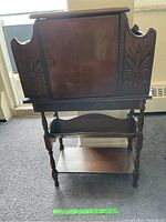 Front view showing the top cabinet with carved floral panels, cupboard door,sidetables, and bottom table with turned legs and diagonal shelf.
