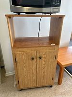 Front view of particle board microwave stand with wood grain finish, upper surface holding a microwave, open middle shelf, and closed lower cabinet with two doors, black knobs, and caster wheels.