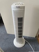 Front view of white Pelonis tower fan standing upright on carpet, showing grille and power cord.