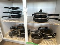 Photo showing wire shelf holding several frying pans with black handles along with a stack of pots and pans beneath including T-fal and other brands, some with glass lids