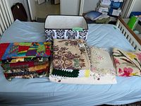 Overview photo showing folded quilts, the hooked tapestry, crocheted blanket, embroidered coverlet, and cloth storage bin on a bed.