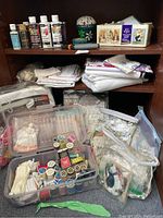 Shelf unit with acrylic paints, folded fabrics, embroidery kits, plastic cases with spools of thread, and small homemade pin cushion