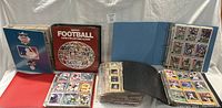 Four binders or albums filled with baseball and football cards shown closed and open to display cards in plastic sleeves, along with loose cards organized on surface.