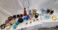 All decorative items arranged on white cloth, including escargot baking dishes, Anthropologie vases, ceramic candle holders, Murano glass dishes, small plant pots, and decorative glass pieces.