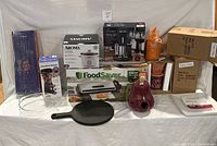 Full lot of mixed kitchen and household items: boxed appliance sets, glassware, cast iron skillet, decorative pitchers, and storage containers.