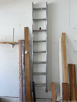 Full view of the 16-foot aluminum ladder mounted against a white wall, surrounded by wooden planks and other objects on the floor and wall.